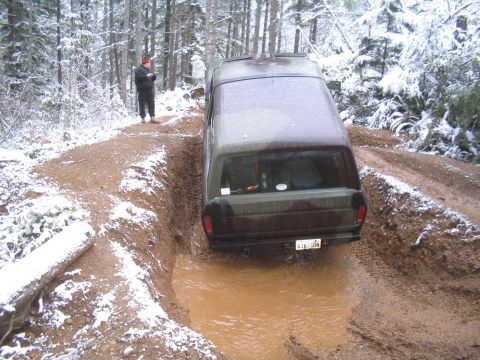 1989 Ford Bronco - A camouflage Ford Bronco is stuck in a muddy rut on a snow-covered dirt road in a forest. A person in a red hat stands on the side of the r…