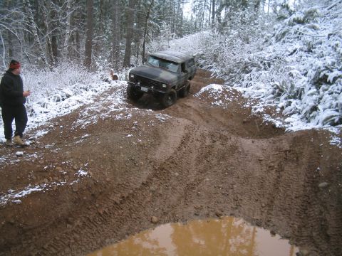1989 Ford Bronco - A camouflaged Ford Bronco is navigating a muddy, snow-covered trail in a wooded area. A person stands on the side of the trail watching the…