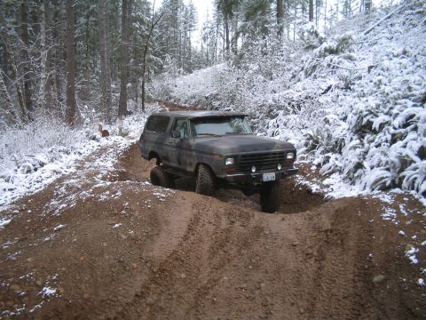 1989 Ford Bronco - A camouflage-painted Ford Bronco is navigating a muddy, rutted trail in a snowy forest. The vehicle is angled towards the viewer, with its…