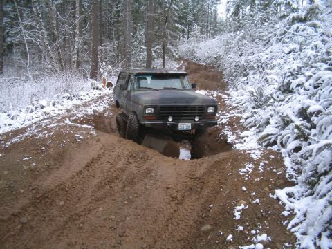 1989 Ford Bronco - A camouflage-painted Ford Bronco navigates a muddy, snow-covered trail through a forest.