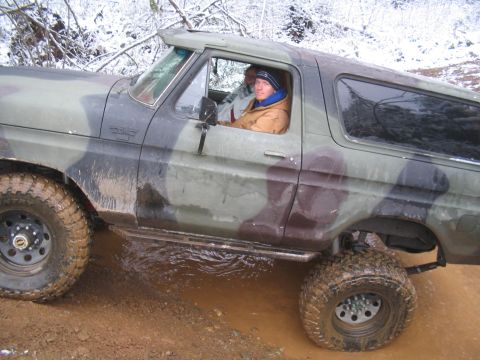 1989 Ford Bronco - A camouflage-painted Ford Bronco is shown driving through muddy water and snow-covered trees are visible in the background.