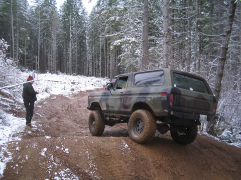 1989 Ford Bronco - A camouflage-painted Ford Bronco is driving off-road on a muddy trail in a snowy forest. A person in a black jacket and beanie stands on th…