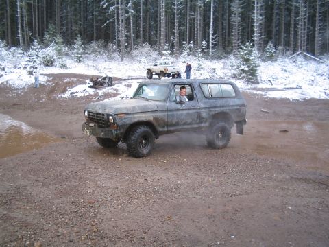1989 Ford Bronco - A weathered, dark-colored Ford Bronco is parked on a muddy, gravelly road in a snowy forest. The driver is visible through the window, smil…