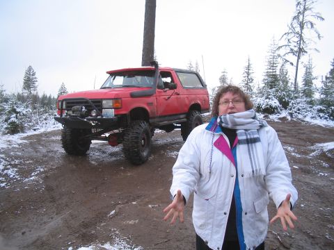 1989 Ford Bronco - A red Ford Bronco with large tires and a snorkel sits on a muddy, snow-dusted trail. A woman stands in front of the vehicle, looking at the…