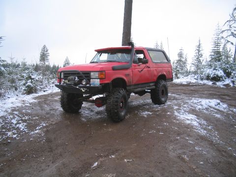 1989 Ford Bronco - A red 1989 Ford Bronco with off-road modifications sits on a muddy, snow-dusted trail in a forest.