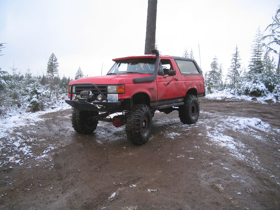 Red 1989 Ford Bronco with off-road tires and winch in a snowy forest.