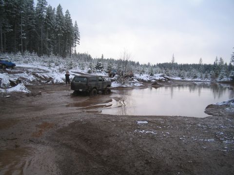 1989 Ford Bronco - A dark-colored SUV is stuck in a muddy mire with snow-covered trees in the background. Another blue vehicle is visible further up a muddy i…