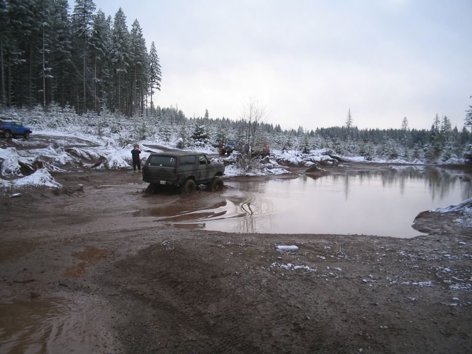 Dark SUV stuck in mud with snow-covered trees and a blue vehicle in the background.