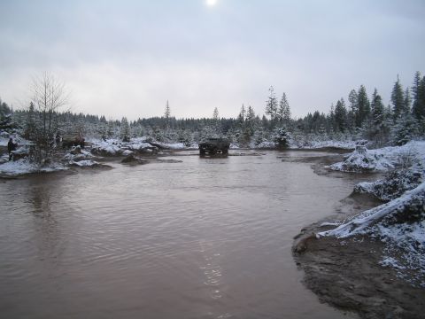 1989 Ford Bronco - A dark-colored SUV drives through a muddy, shallow body of water surrounded by snow-covered trees. Another red SUV is parked on the bank in…