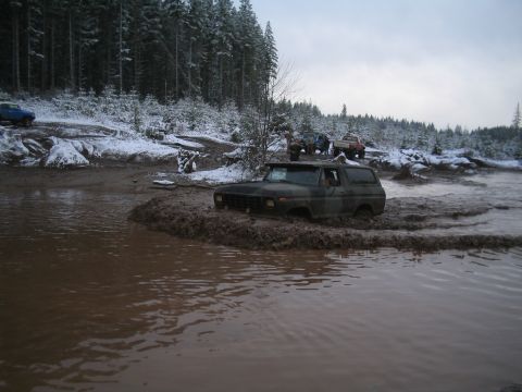 1989 Ford Bronco - A dark-colored SUV is stuck deep in thick mud with snow on the ground and trees in the background. Several other off-road vehicles are visi…