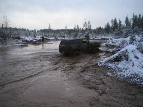 1989 Ford Bronco - A dark-colored Ford Bronco is navigating a muddy, water-filled off-road trail with snow-covered trees in the background.