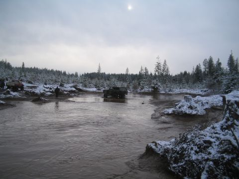 1989 Ford Bronco - A dark-colored SUV is driving through a muddy, shallow body of water with snow-covered trees and ground in the background. Another red pick…
