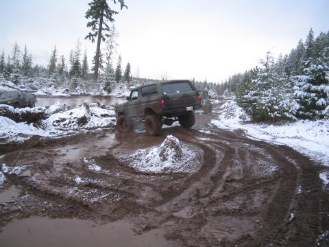 1989 Ford Bronco - A camouflaged Ford Bronco navigates a muddy, snow-covered trail with a river and dense forest in the background. Another vehicle is visible…