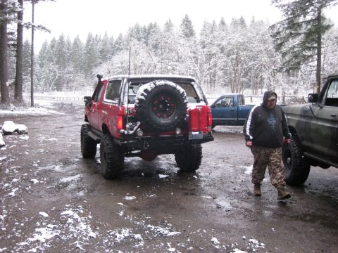 1989 Ford Bronco - A red Ford Bronco with a spare tire mounted on the rear is parked in a muddy, snow-dusted lot next to a dark green pickup truck. A man wear…