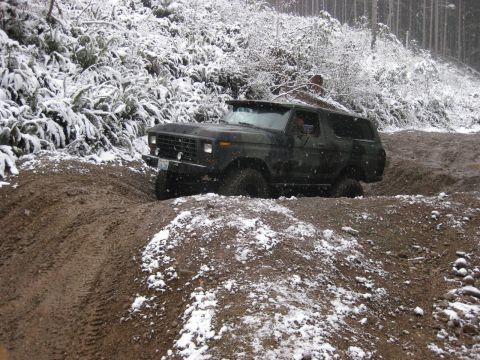 1989 Ford Bronco - A dark-colored Ford Bronco is navigating a muddy, snow-dusted off-road trail. The vehicle is angled to the right, with its front tire deep…