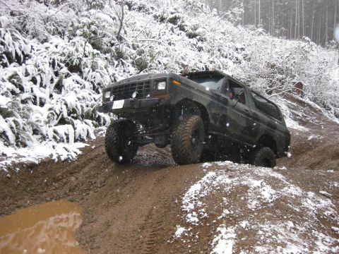 1989 Ford Bronco - A dark colored Ford Bronco is navigating a muddy, snow-covered trail, with its tires deep in the mud and snow.