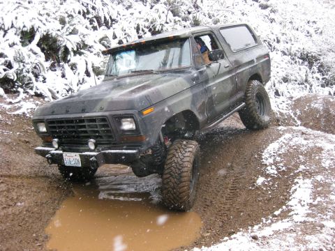 1989 Ford Bronco - A camouflage Ford Bronco is driving through a muddy puddle with snow falling around it. The vehicle is on an off-road trail with snow-cover…