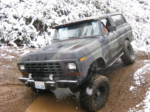 1989 Ford Bronco - A camouflage Ford Bronco is driving off-road through mud and snow, with its front tires deep in a muddy puddle. The vehicle has large off-r…