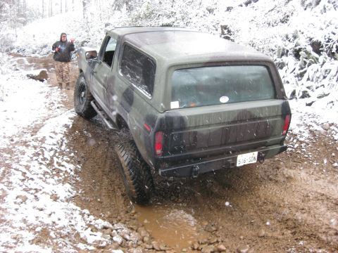 1989 Ford Bronco - A camouflage-painted Ford Bronco is stuck in mud on a snowy, wooded trail. The driver stands beside the vehicle, giving a thumbs-up.