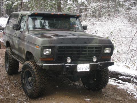 1989 Ford Bronco - A camouflage-painted Ford Bronco with large off-road tires is parked on a muddy, snow-dusted trail in a forest. The front of the vehicle is…