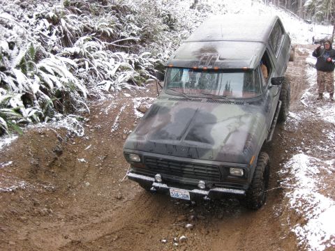 1989 Ford Bronco - A camouflage-painted Ford Bronco is driving on a muddy, snow-dusted trail with dense foliage on the left. A person stands on the right side…