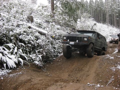 1989 Ford Bronco - A dark-colored, lifted Ford Bronco is driving off-road on a muddy trail covered in patches of snow. Another off-road vehicle is visible in…