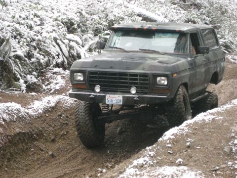 1989 Ford Bronco - A dark colored, lifted 1980s Ford Bronco is navigating a muddy, snow-dusted trail. The vehicle is angled to the right, showing its front an…