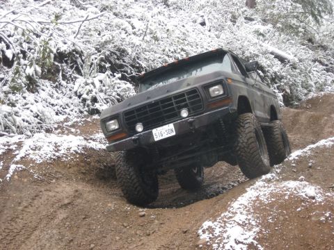 1989 Ford Bronco - A lifted, dark-colored 4x4 truck with large tires navigates a muddy, snow-dusted trail. The vehicle is angled uphill, showing its front-lef…