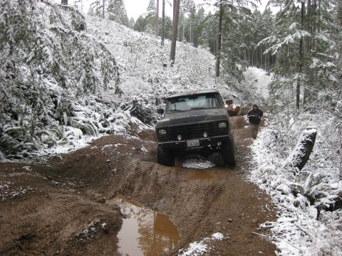 1989 Ford Bronco - A black Ford Bronco navigates a muddy and snow-covered off-road trail in a forest. Another SUV is visible further up the trail, with a pers…