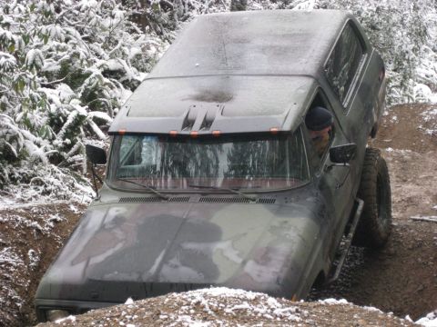 1989 Ford Bronco - A camouflage-painted SUV is navigating a muddy, rutted path with snow-covered trees in the background. The driver, wearing a dark beanie, i…