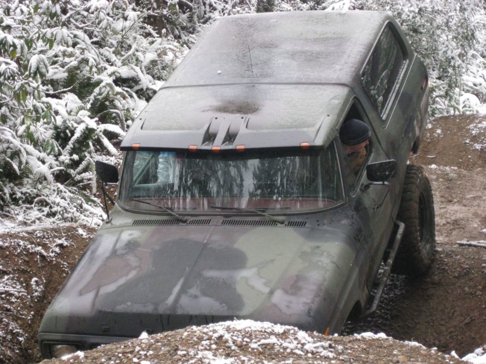 Camouflage SUV driving through a muddy rut with snow on surrounding trees.