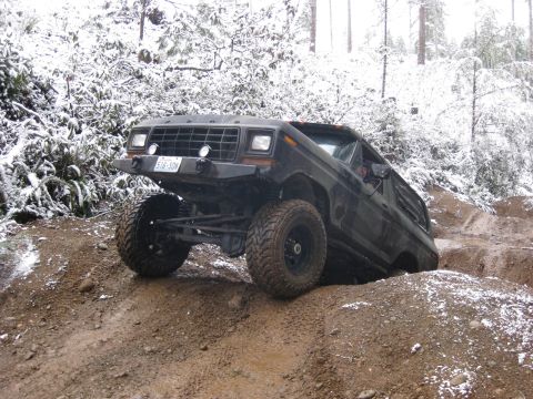 1989 Ford Bronco - A dark-colored, lifted Ford Bronco is navigating a muddy, off-road trail with snow-covered trees in the background. The vehicle is angled o…