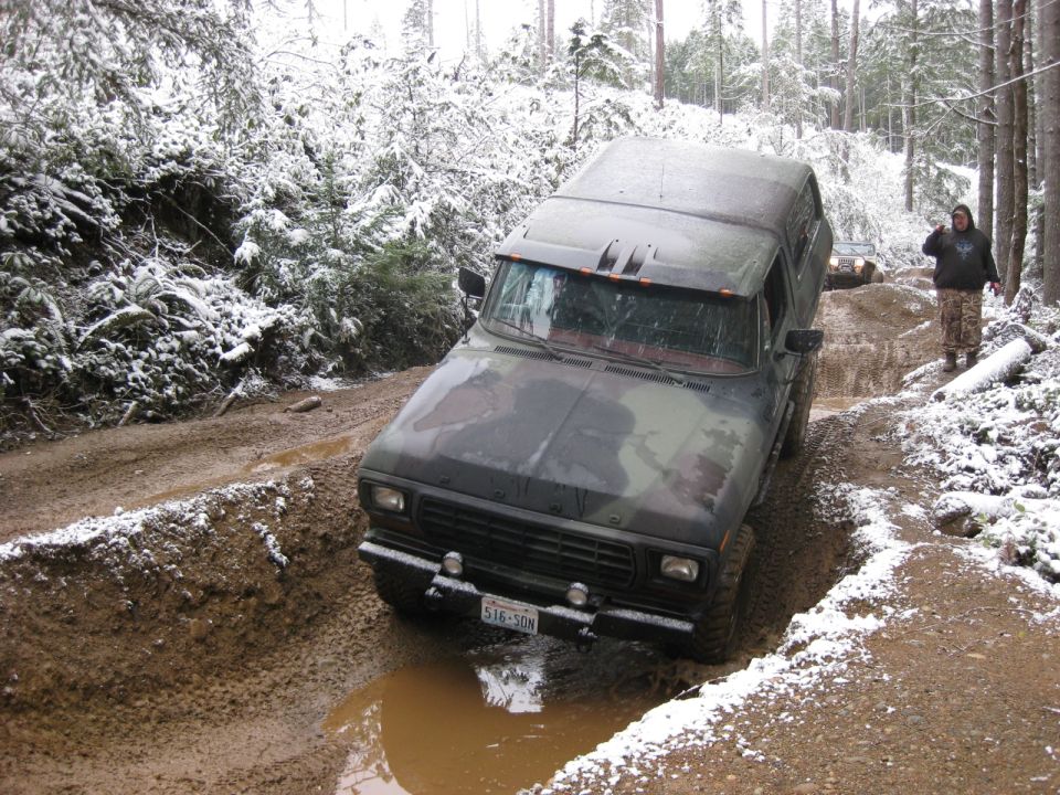 Camouflage Ford Bronco navigating a muddy, snow-covered forest road.