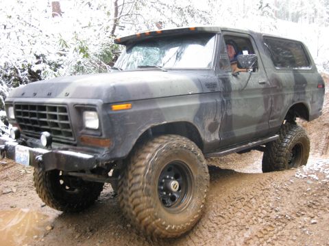 1989 Ford Bronco - A camouflage-painted Ford Bronco is driving through a muddy, snowy terrain. The vehicle is angled to the right, showing its front and side…