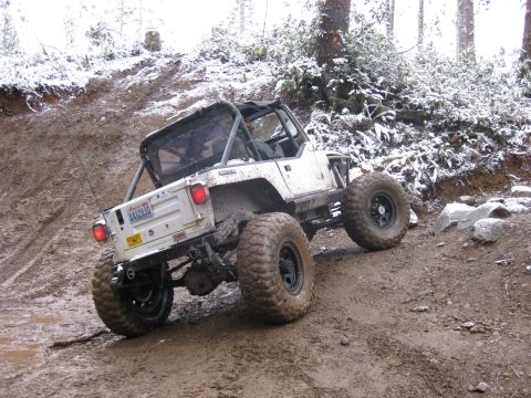 1989 Ford Bronco - A white Jeep Wrangler is navigating a muddy, snow-dusted off-road trail. The vehicle is angled upwards on a steep incline, with large, knob…