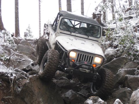 1989 Ford Bronco - A white Jeep Wrangler is navigating a rocky, snow-covered trail with large off-road tires. The vehicle is angled upwards, climbing over bou…
