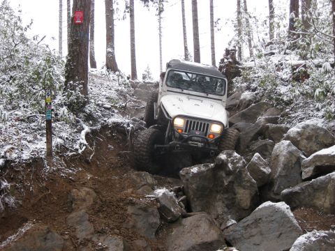 1989 Ford Bronco - A white Jeep Wrangler is navigating a rocky, snow-covered trail in a forest. The vehicle is angled towards the camera, with its front tires…
