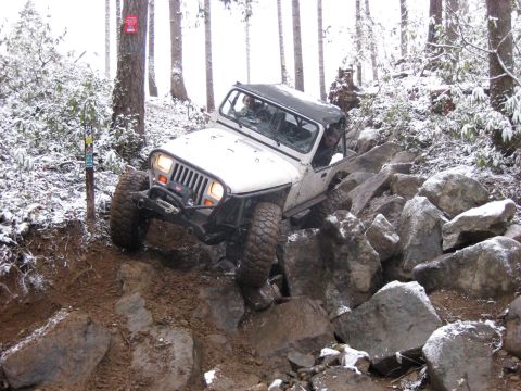 1989 Ford Bronco - A white Jeep Wrangler is navigating a rocky, muddy trail in a snowy forest. The vehicle is angled upwards, with its front tires on large ro…