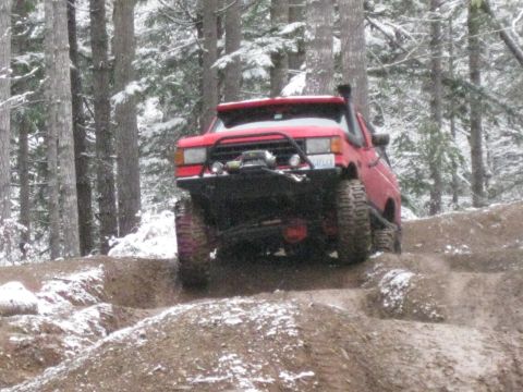 1989 Ford Bronco - A red off-road SUV with large tires navigates a muddy, snow-dusted trail through a pine forest.
