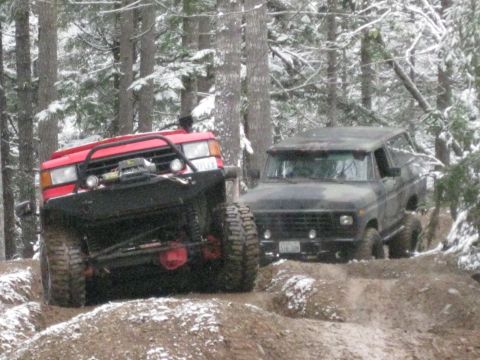 1989 Ford Bronco - Two off-road vehicles are navigating a muddy trail in a snowy forest. The vehicle in the foreground is red with a winch and custom bumper,…