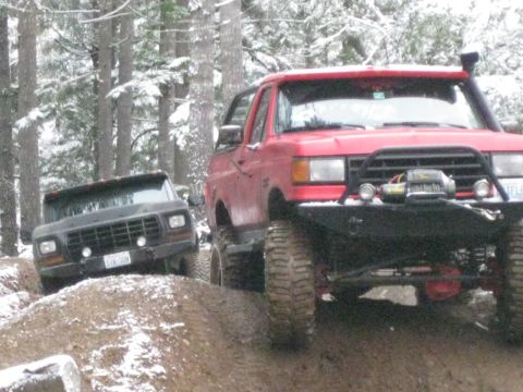 1989 Ford Bronco - Two off-road trucks, one red and one black, navigate a muddy, snow-covered trail in a wooded area. The red truck in the foreground features…