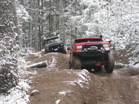 1989 Ford Bronco - Two off-road modified trucks navigate a muddy trail covered in snow in a forest. The truck in the foreground is red with large tires and a…