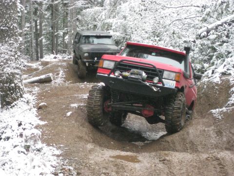 1989 Ford Bronco - Two off-road vehicles navigate a muddy, snow-dusted trail through a forest. The vehicle in the foreground is a red, lifted truck with large…