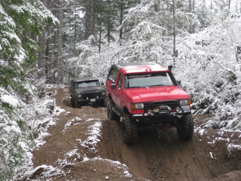 1989 Ford Bronco - Two off-road vehicles navigate a muddy, snow-covered forest trail. The lead vehicle is a red SUV, and the second vehicle behind it is a dar…