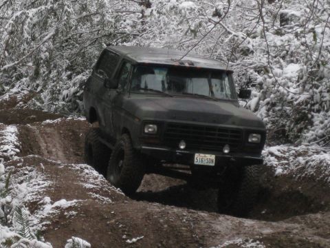 1989 Ford Bronco - A dark colored, lifted Ford Bronco is shown driving off-road on a muddy trail with snow on the surrounding trees and ground. The vehicle's…