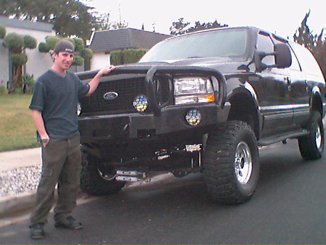 Lifted black Ford Excursion with brush guard bumper and big off-road tires; owner standing beside it on a street