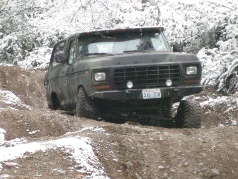 1989 Ford Bronco - A dark colored Ford Bronco is driving on a dirt path with snow on the ground and trees in the background. The vehicle has a license plate t…