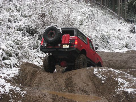 1989 Ford Bronco - A red 1989 Ford Bronco is shown from the rear 3/4 view, navigating a muddy, snow-covered off-road trail. The vehicle has a spare tire mount…