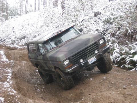 1989 Ford Bronco - A camouflage-painted 1980s Ford Bronco is navigating a muddy, snow-dusted off-road trail. The vehicle is angled sharply to its right, with…