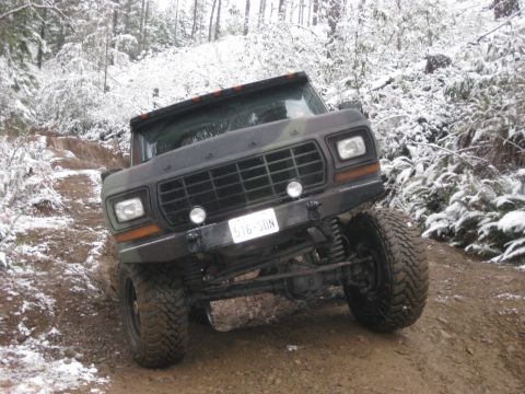 1989 Ford Bronco - A dark-colored, lifted Ford Bronco is navigating a muddy, snow-covered trail in a wooded area. The front of the vehicle is angled towards t…